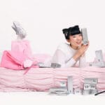 Una modelo asiática posa tumbada en una cama rosa con las cajitas del calendario de adviento MiiN Cosmetics 2025