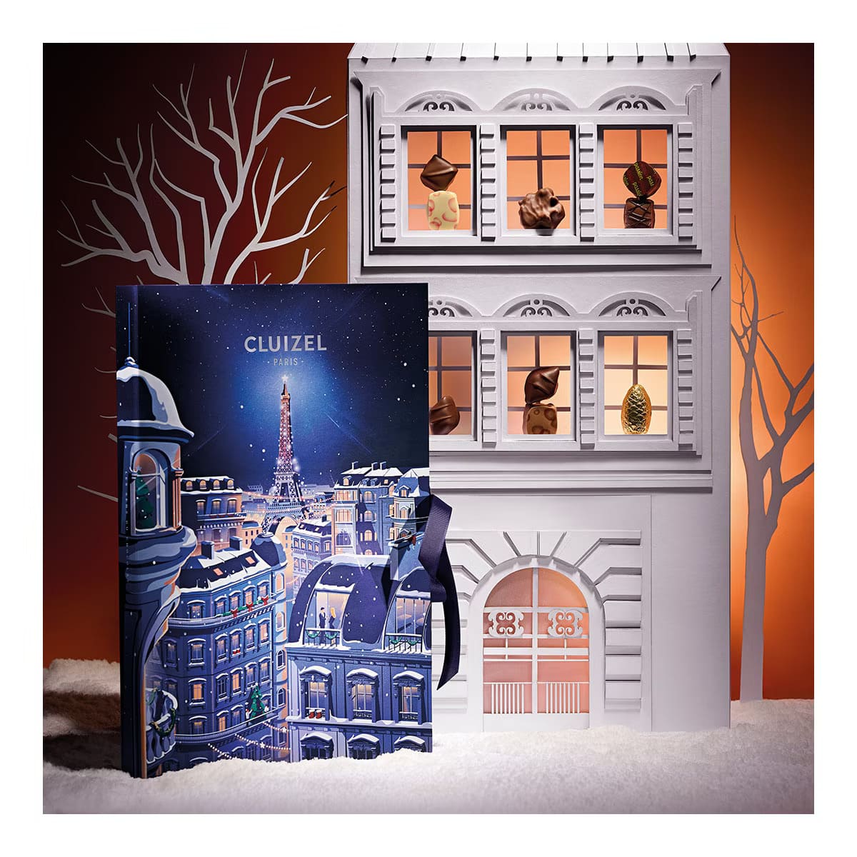 La caja del Calendario de Adviento Michel Cluizel 2023 junto a la maqueta de una fachada en cuyas ventanas hay bombones de chocolate