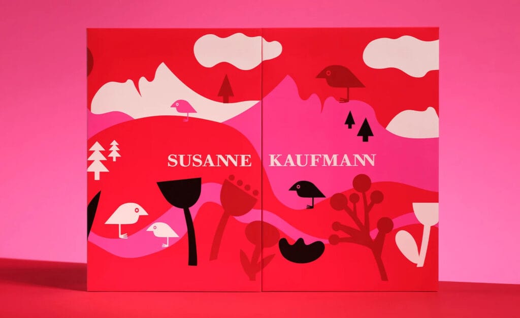 La parte frontal de la caja del Calendario de adviento Susanne Kaufmann 2025, en tonos rojos, rosas y blancos y con el logo del ave de la marca en un paisaje de montañas y vegetación