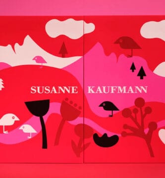La parte frontal de la caja del Calendario de adviento Susanne Kaufmann 2025, en tonos rojos, rosas y blancos y con el logo del ave de la marca en un paisaje de montañas y vegetación