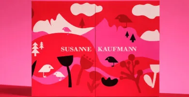La parte frontal de la caja del Calendario de adviento Susanne Kaufmann 2025, en tonos rojos, rosas y blancos y con el logo del ave de la marca en un paisaje de montañas y vegetación